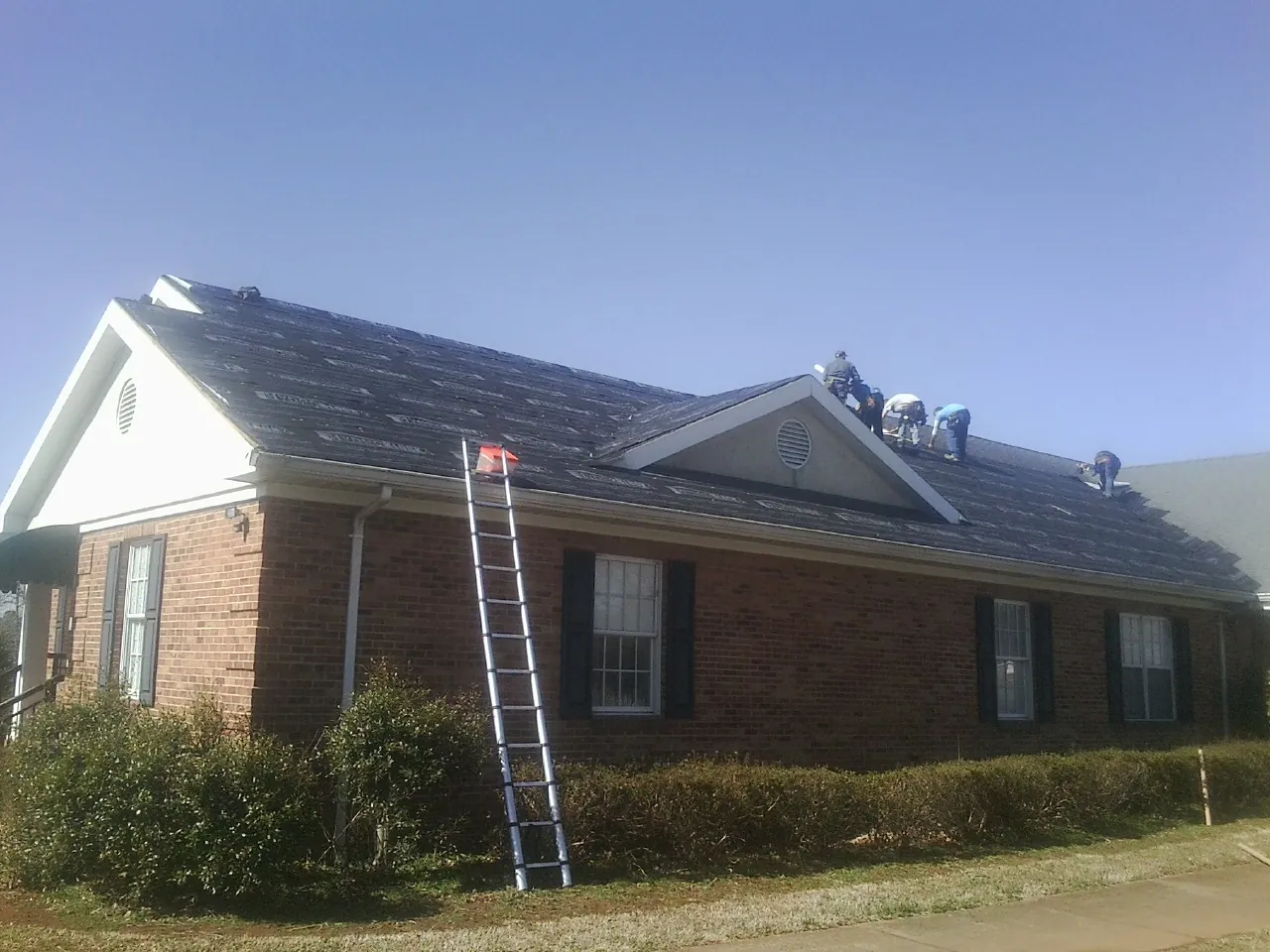 Crew tearing shingles off a brick ranch roof with a ladder set — Emergency Roof Repair work by our crew in North Billerica