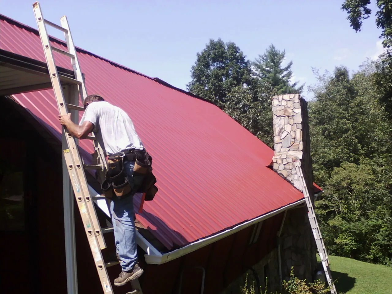 Roofer on a ladder beside a red metal roof and stone chimney — Soffit & Fascia Repair by Roofing Brothers in North Billerica